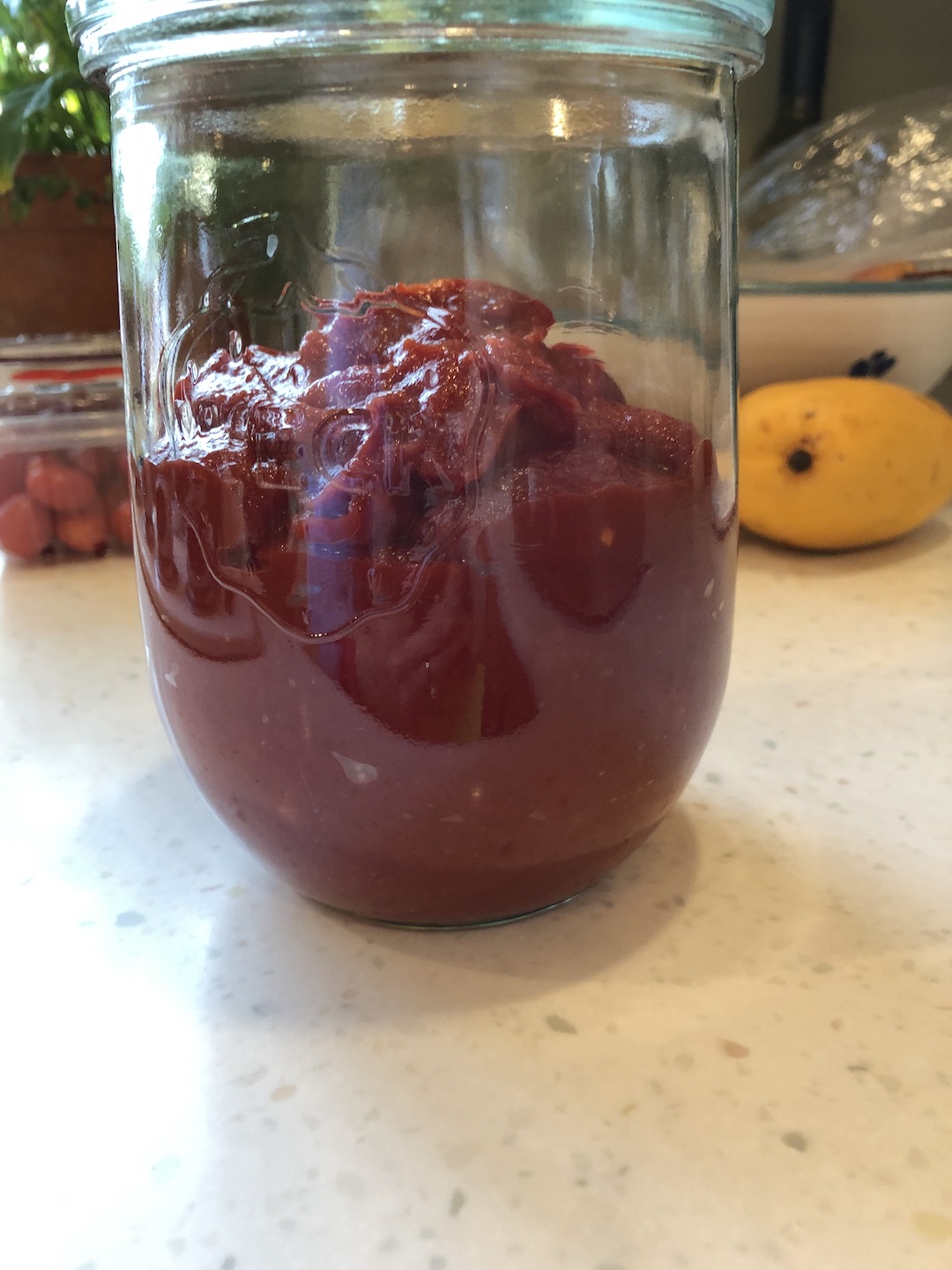 ketchup sitting in a glass jar on the counter fermenting with a mango in the background