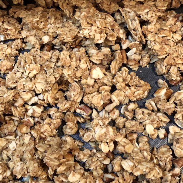 granola with pecans in chunks sitting on a cookie sheet with a black liner
