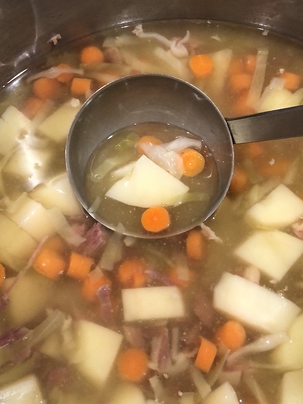 ladle with carrots, potato and cabbage pieces in a ladel with broth over a pot of soup