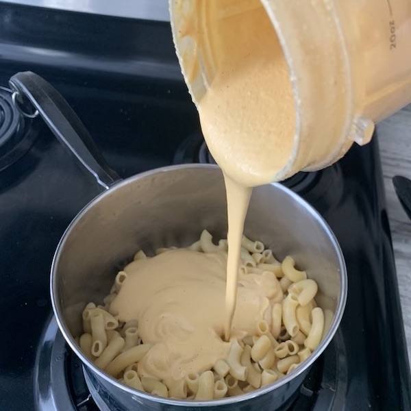 Pouring pale orange sauce from blender container into pot with drained noodles.