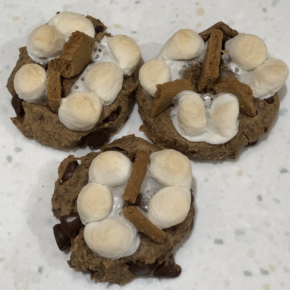 Three vegan s'mores cookies with mini marshmallows toasted on top sitting on a white speckled counter.