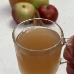 apple peel tea in clear mug with apples, cinnamon sticks and carafe of tea in background