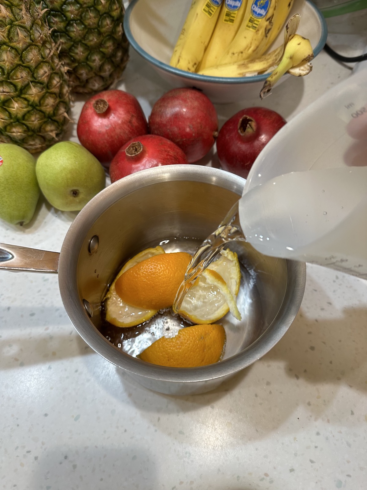 Orange peel in a small pot with water being poured in. Pomegranates, pears, pineapple and bananas in the background.