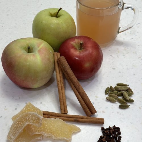 apples, cinnamon sticks, crystoalized ginger, whole cloves, cardamom pods and a cup of apple tea in a clear mug