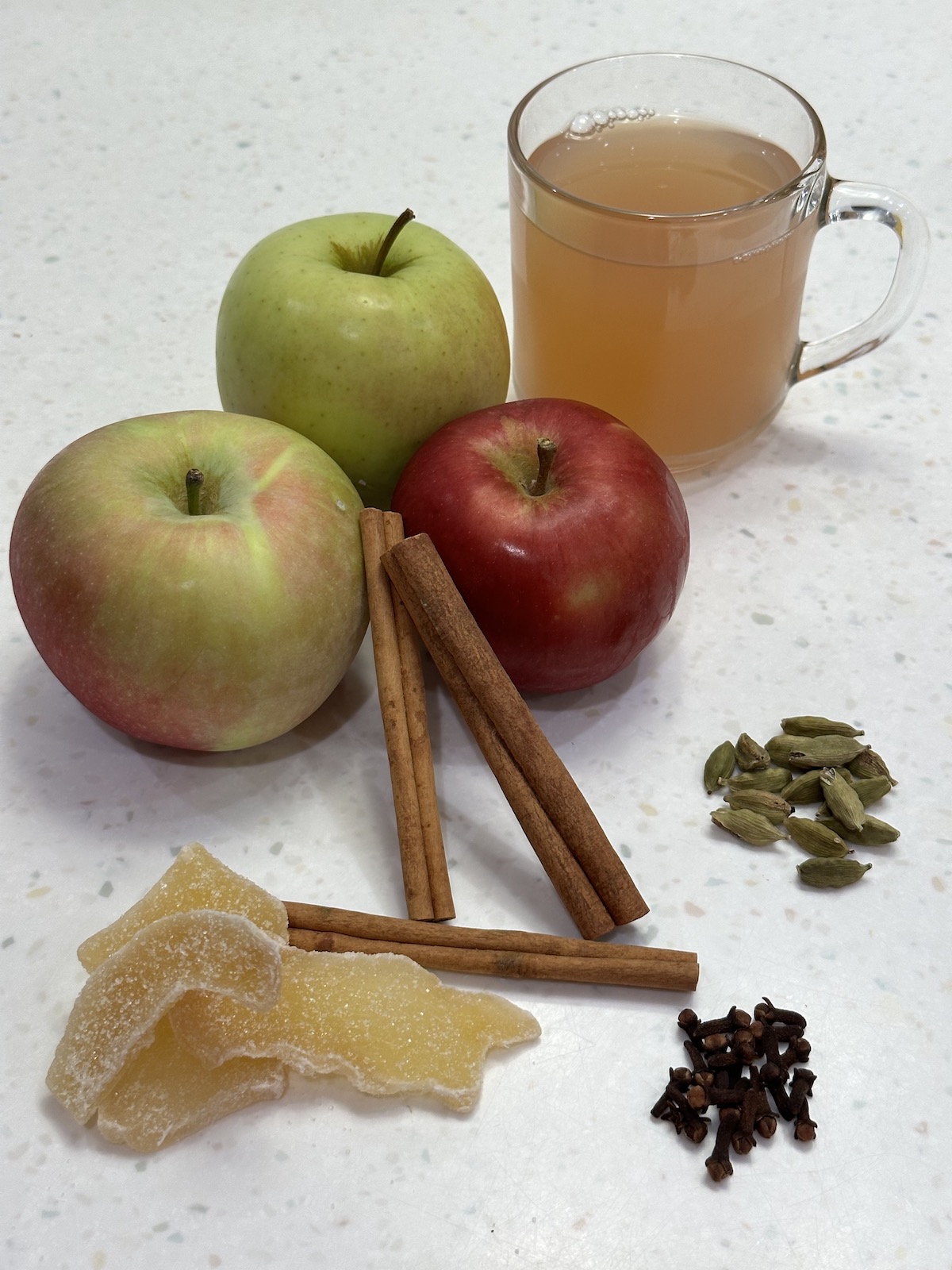 apples, cinnamon sticks, crystoalized ginger, whole cloves, cardamom pods and a cup of apple tea in a clear mug