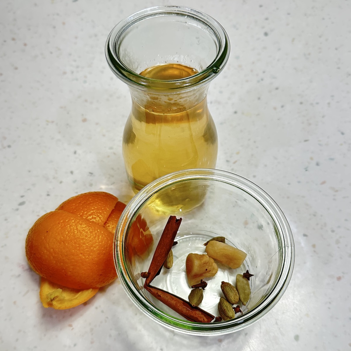 small carafe of orange peel tea with an orange peel by the side and spices in a clear open jar