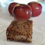 A square piece of apple cake sitting on a glass plate with three red apples behind.