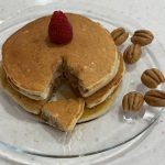 3 pancakes stacked with pecans on a clear plate and a raspberry on top. Pancake stack has a triangle cut out.